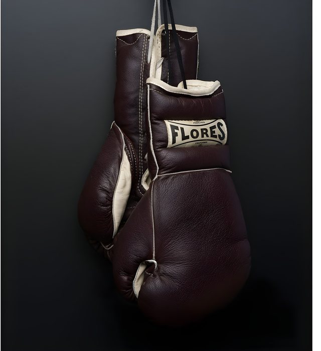 A pair of brown leather FLORES gloves hanging by their laces against a dark charcoal ground, the cream FLORES San Francisco California cuff label facing the camera.